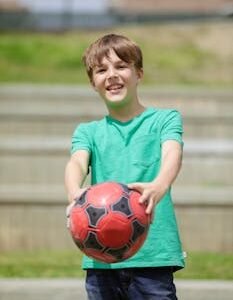 Young boy smiling, holding a soccer ball outdoors with blurred steps background